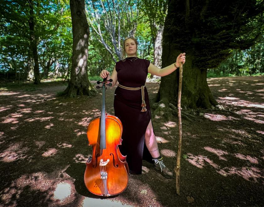 Non-binary person in a red dress standing in a forest holding a cello