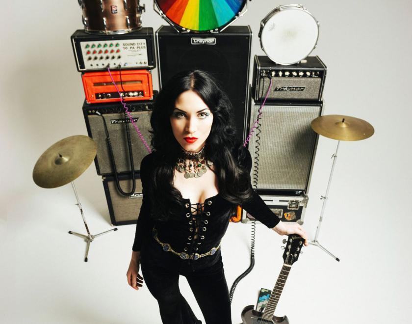 Rosalie Cunningham wearing a black velvet jupsuit standing in front of a stack of amps and instruments
