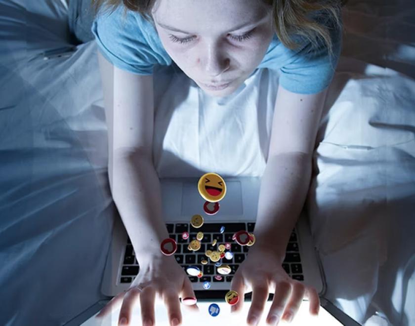 Young girl in bed on her laptop. Various emojis appear to be spilling out of the screen into her hands.