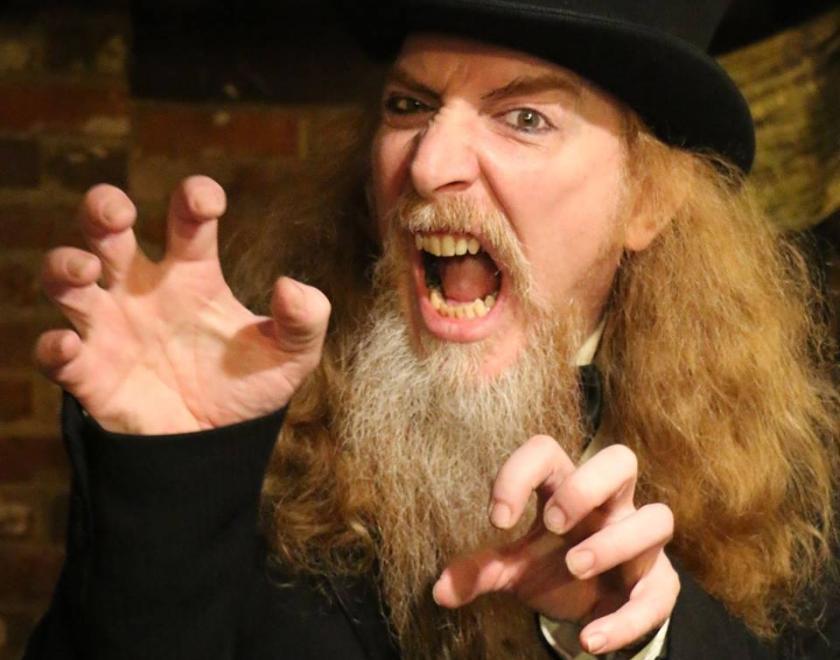 Long haired ginger chap with a grey beard wearing a top hat and black coat growls at the camera