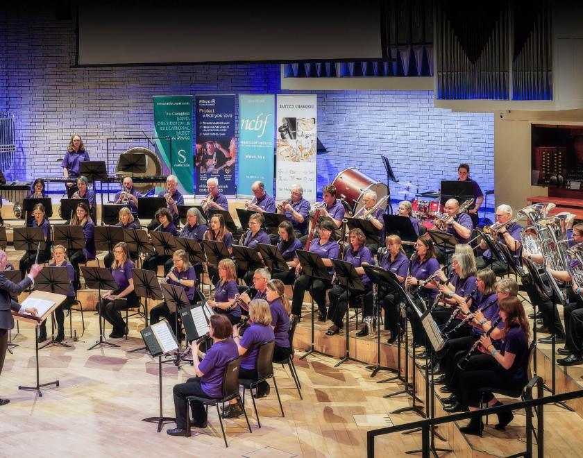 Trinity Concert Band performing in a festival, with the conductor leading the large seated wind band on stage.