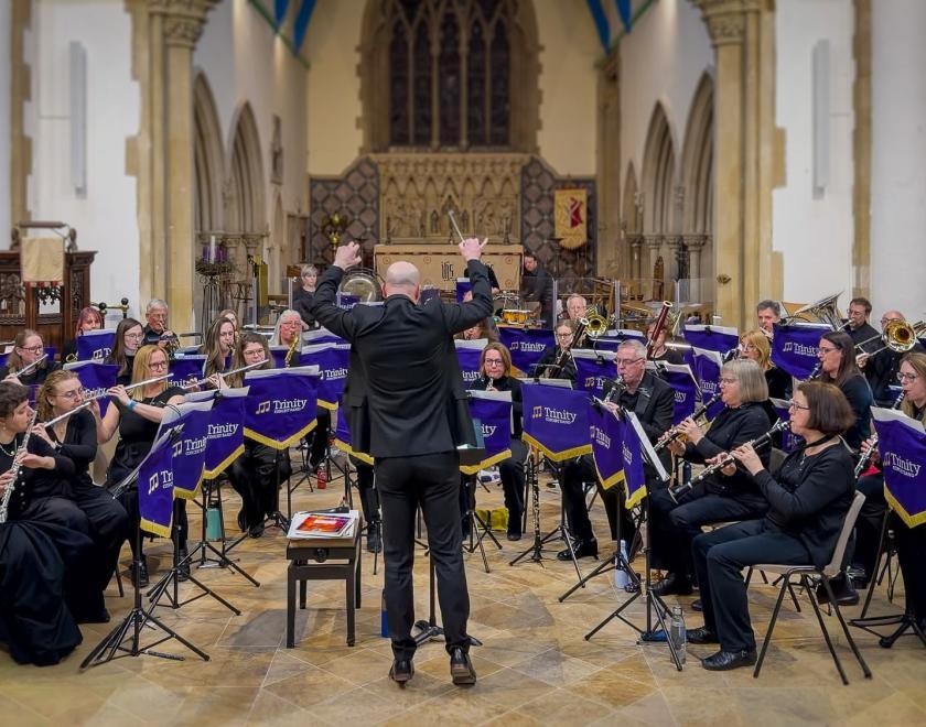 Trinity Concert Band performing in a church, with the conductor leading the seated wind band from the centre.