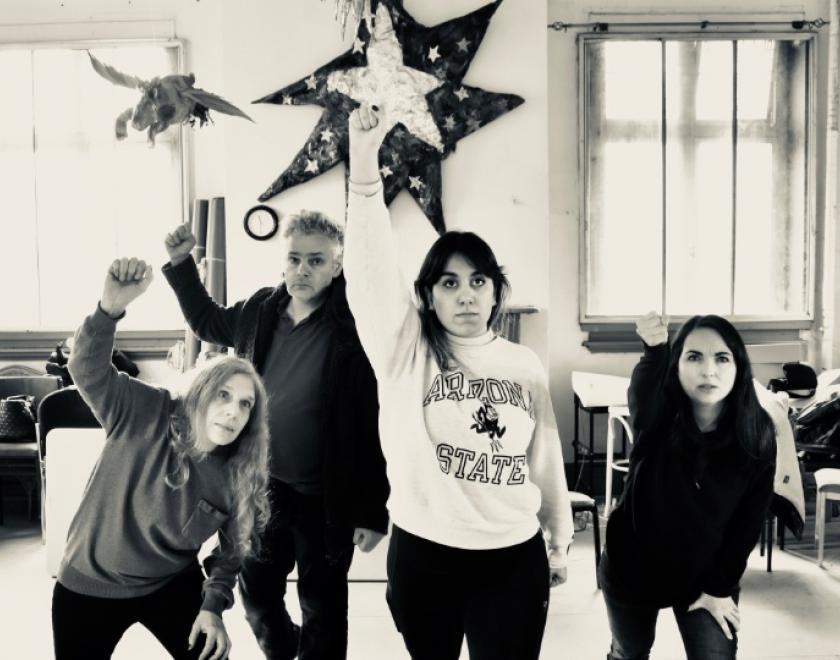 A black and white photo, three women and a man stand with one arm raised in a fist, they look seriously into the camera