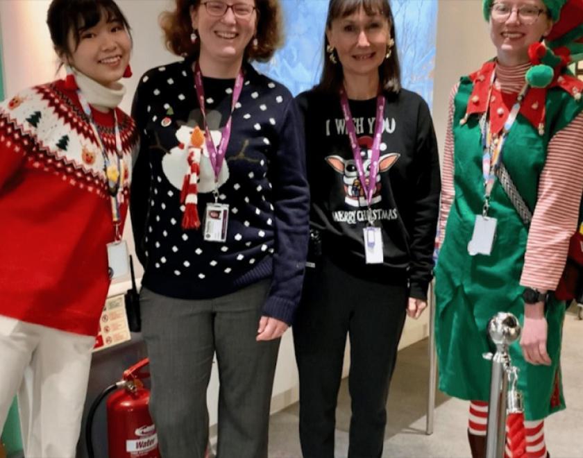 three women wearing Christmas jumpers, and a fourth dressed as an elf, standing in a line and smiling